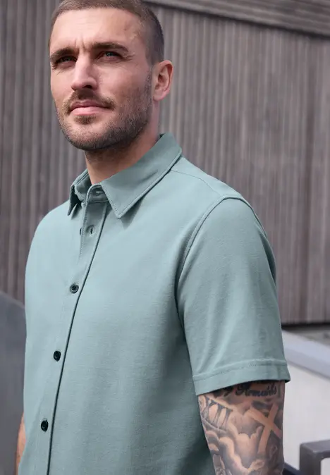 Un homme en chemise verte pâle à manches courtes avec des bras tatoués devant un mur en bois.