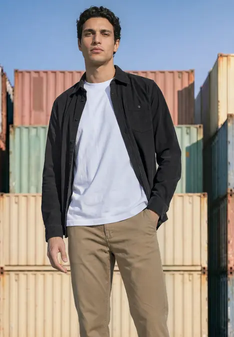 Ein Mann in weißem T-Shirt, schwarzer Jacke und khakifarbenen Hosen steht vor bunten Containern im Freien.