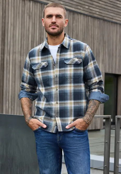Ein bärtiger Mann in karierter Flanelljacke und Jeans lehnt an einem Geländer vor einer Holzfassade.