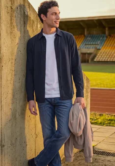 Ein junger Mann lehnt entspannt an einer Betonwand, trägt Jacke, Jeans und T-Shirt vor einer Tribüne.