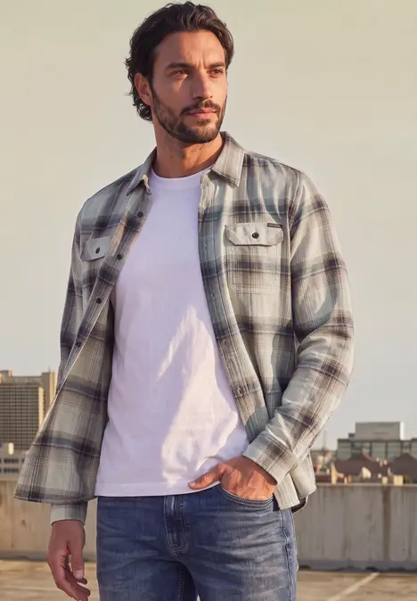 Ein Mann in kariertem Hemd und weißem T-Shirt steht vor urbaner Kulisse und schaut nach rechts.