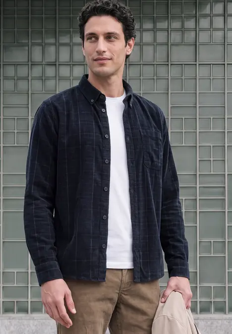 Un jeune homme se tient devant un mur de carreaux de verre, vêtu d’un t-shirt blanc, d’une chemise à carreaux ouverte et d’un pantalon marron.