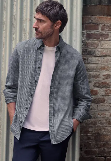 Un homme barbu en chemise grise et tee-shirt rose se tient contre un mur en regardant sur le côté.