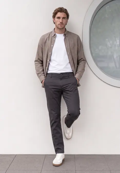 Un homme est appuyé contre un mur, vêtu d’un t-shirt blanc, d’un pantalon gris, d’une veste brune et de baskets blanches.