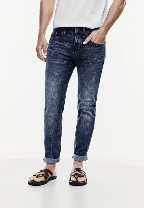 Man draagt spijkerbroek, wit shirt en sandalen.
