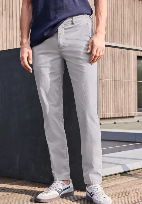 Homme en pantalon chino gris et baskets, vue du bas du corps devant un mur en bois