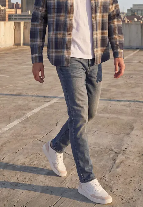 Een man in jeans, wit T-shirt, geruit overhemd en witte sneakers op een betonnen parkeerdek.