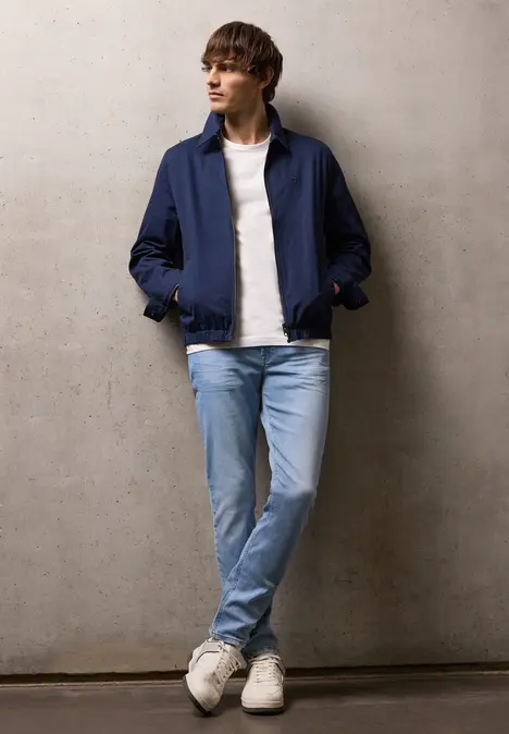 Een persoon staat ontspannen voor een grijze muur, met een blauwe jas, wit shirt, lichte jeans en witte sneakers.