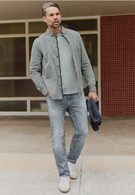 Een man in een casual denimoutfit (jeans, overhemd, T-shirt) met sneakers, houdend een jas voor een gebouw.