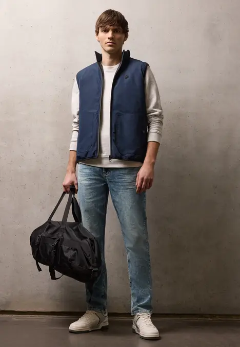 Man in blauwe bodywarmer, grijs sweatshirt, jeans en witte sneakers met een zwarte reistas.