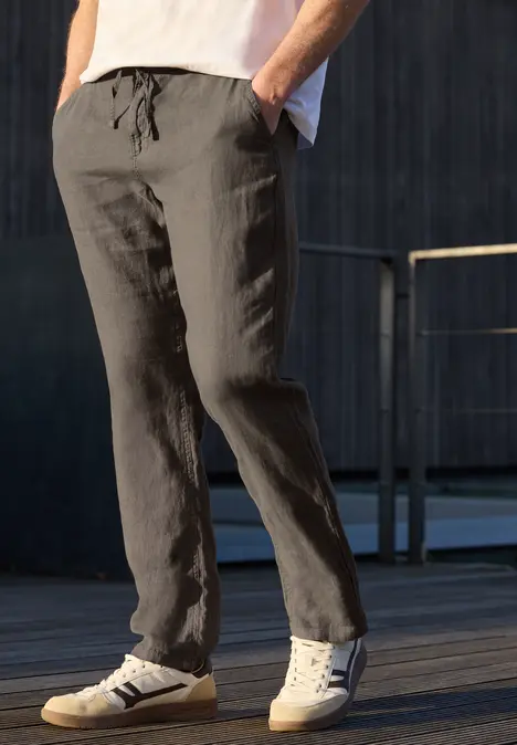 Homme en pantalon gris décontracté et baskets blanches, debout sur un sol en bois en extérieur.
