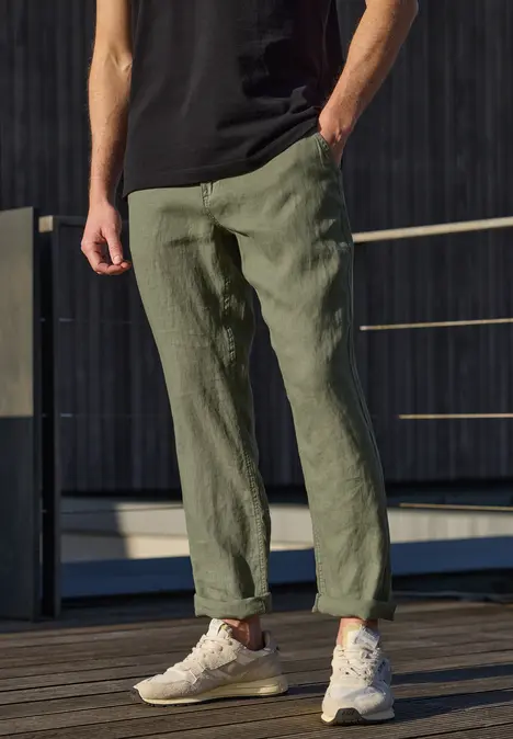 Un homme élancé se tient sur une terrasse en bois, porte un pantalon vert en lin, un t-shirt noir et des baskets blanches.