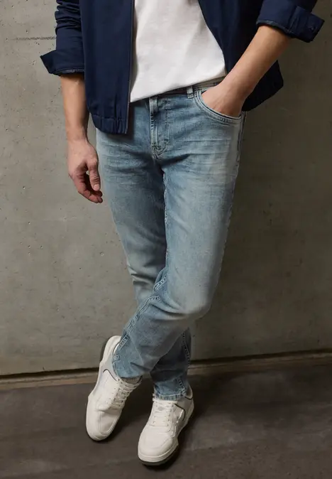 Persoon leunt tegen een grijze muur, draagt witte sneakers, blauwe jeans, wit T-shirt en blauwe jas.