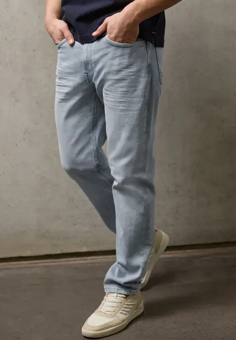Iemand draagt lichtblauwe spijkerbroek en witte sneakers voor een betonnen muur.