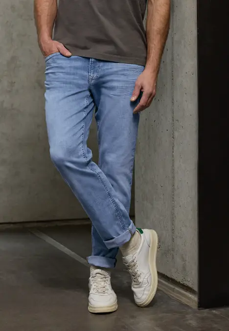 Een man in lichtblauwe jeans en witte sneakers leunt nonchalant tegen een betonnen muur.