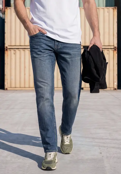 Man in spijkerbroek en wit T-shirt met groene sneakers, draagt zwarte jas en loopt langs containers.