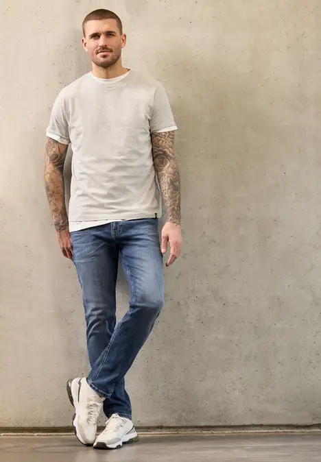Een getatoeëerde man in jeans en wit T-shirt leunt ontspannen tegen een grijze muur.