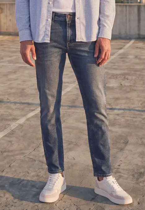 Homme en jean sombre et baskets blanches, debout sur un sol en béton.