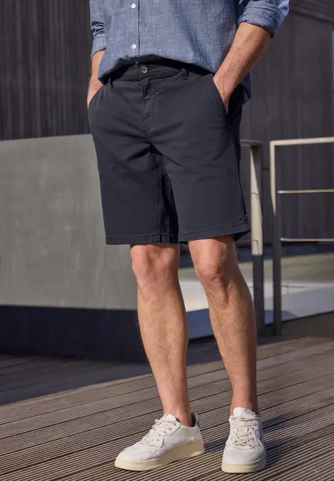 Mann trägt dunkelblaue Shorts, hellblaue Hemd und weiße Sneakers, steht auf Holzterrasse vor modernem Gebäude.