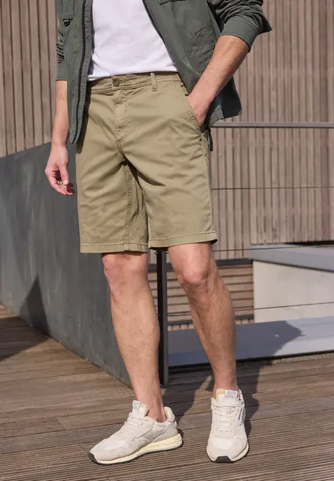 Ein Mann in khakifarbenen Shorts, weißem T-Shirt, grüner Jacke und hellen Sneakern auf Holzterrasse.