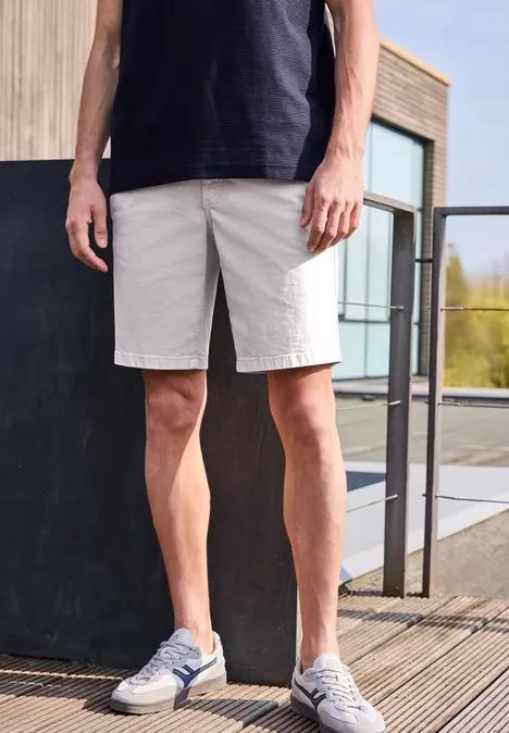 Ein Mann trägt ein schwarzes T-Shirt, graue Shorts und Sneaker, stehend auf einer Holzterrasse vor einem modernen Gebäude.