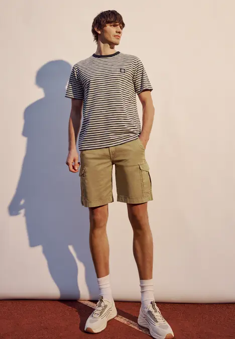 Ein junger Mann steht vor einer hellen Wand, trägt ein gestreiftes T-Shirt, beige Shorts und weiße Sneakers.