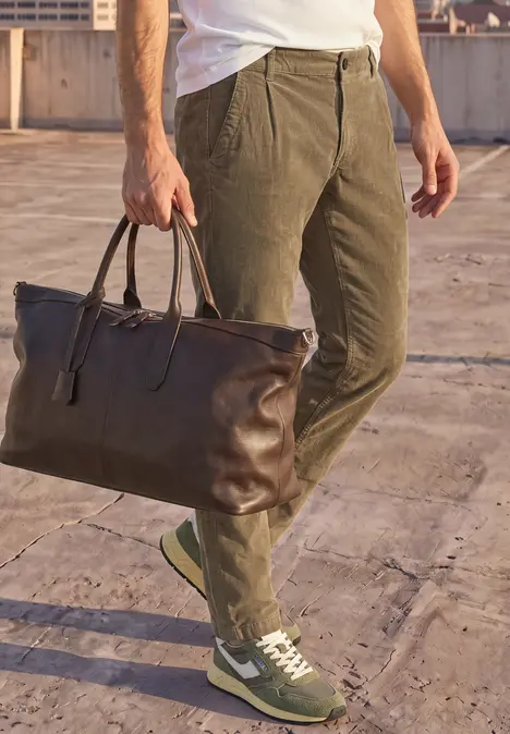 Mann in olivgrüner Hose und Sneakern trägt eine große braune Ledertasche auf einem Dach.