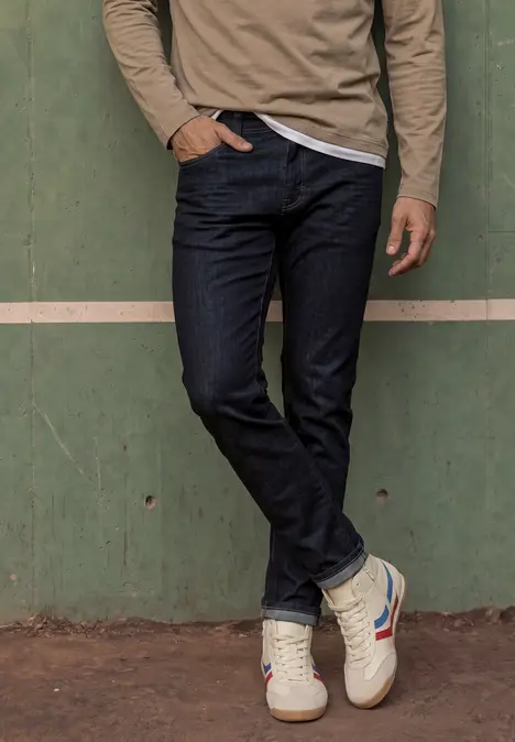 Ein Mann in Jeans und Sneakers steht vor einer grünen Wand, eine Hand in der Hosentasche.