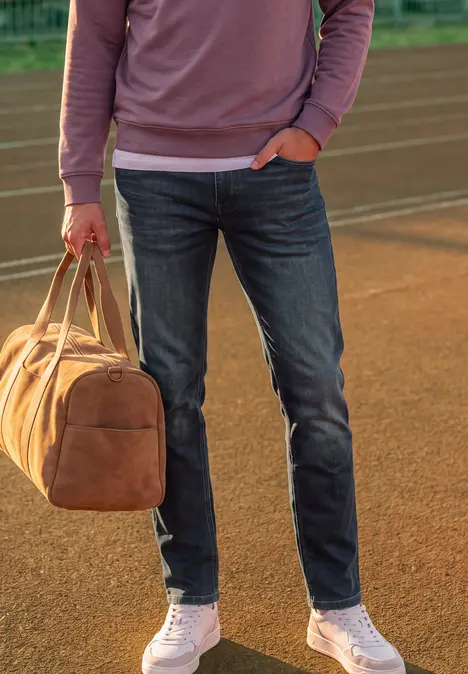 Person in Jeans, weißen Sneakern und rosa Sweatshirt hält braune Reisetasche auf Laufbahn.