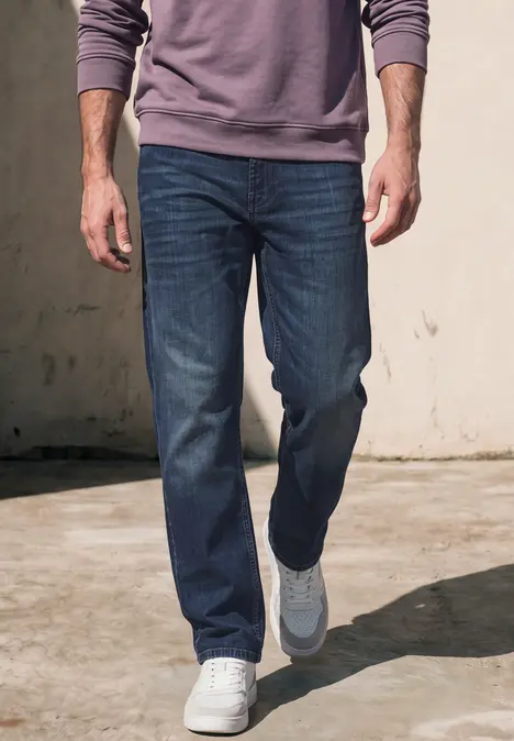 Mann in blauen Jeans und weißen Sneakern geht vor einer hellen Wand.