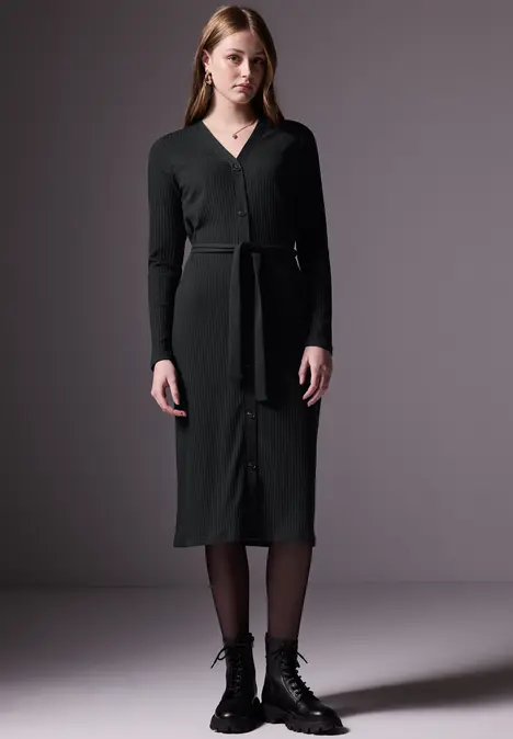 Frau in einem dunkelgrünen Strickkleid mit Gürtel und schwarzen Stiefeln.