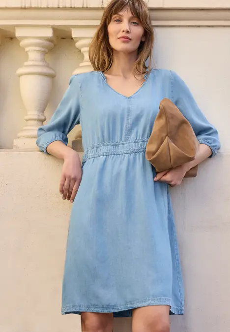 Vrouw in licht denimjurk met V-hals en driekwartmouwen, met een bruine tas in de hand.