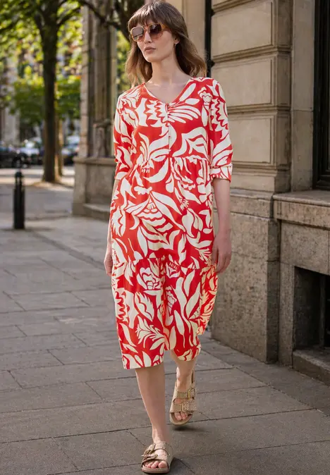 Eine Frau in einem rot-weißen Midikleid mit abstraktem Muster spaziert auf einem Gehweg vor einem Steinbau.
