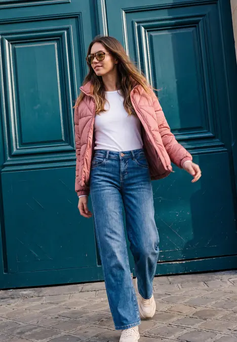 Une femme souriante en veste rose et jean porte des lunettes de soleil devant une porte bleue.