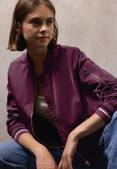 Frau mit kurzer brauner Jacke und weißem Shirt sitzt vor grauer Wand.