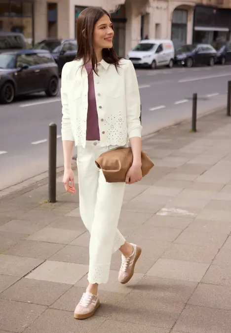 Vrouw in witte denim en bordeaux top, met beige schoudertasje en roségouden sneakers op stoep.