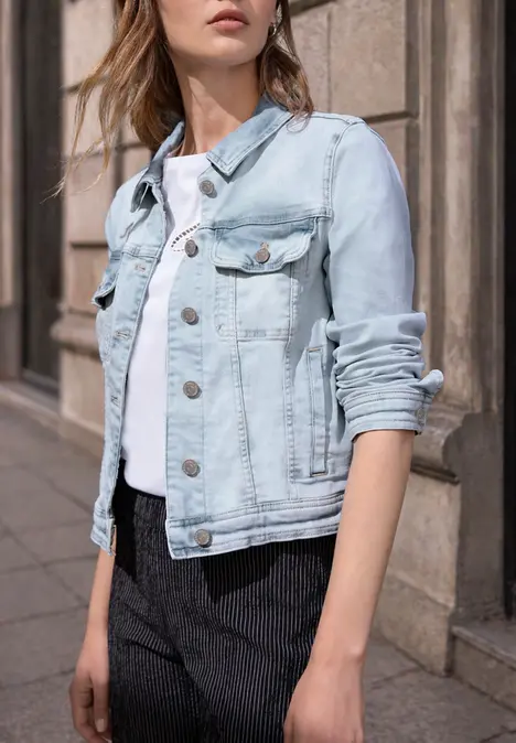 Vrouw in lichtblauw jeansjack, wit T-shirt en zwarte broek, staand in een stedelijke omgeving.