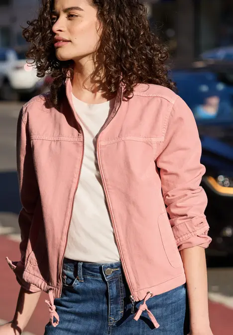 Een persoon met krullend haar draagt een roze jas, wit shirt en spijkerbroek voor een geparkeerde auto.