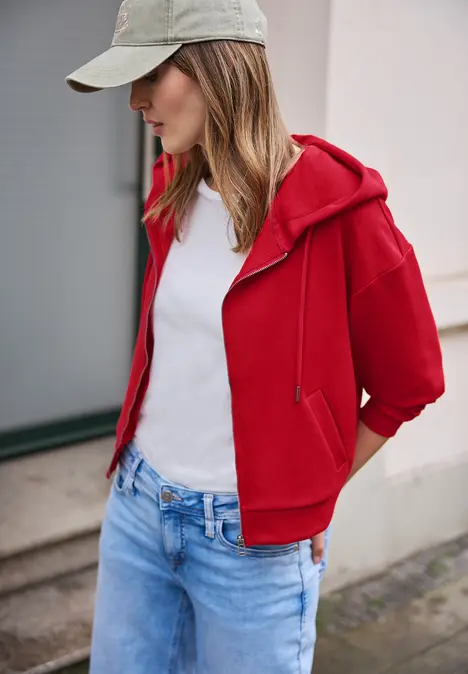 Eine Person in roter Kapuzenjacke, weißem T-Shirt, blauer Jeans und Baseballcap vor einer Hauswand.