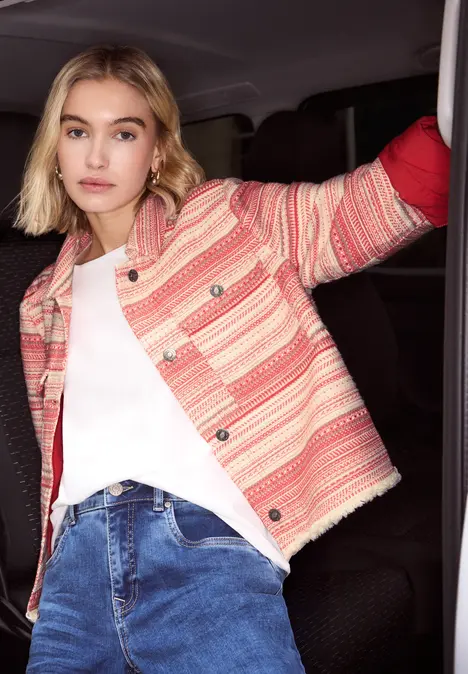 Eine Person trägt eine rosé-gestreifte Jacke, weißes T-Shirt und blaue Jeans im Fahrzeug.