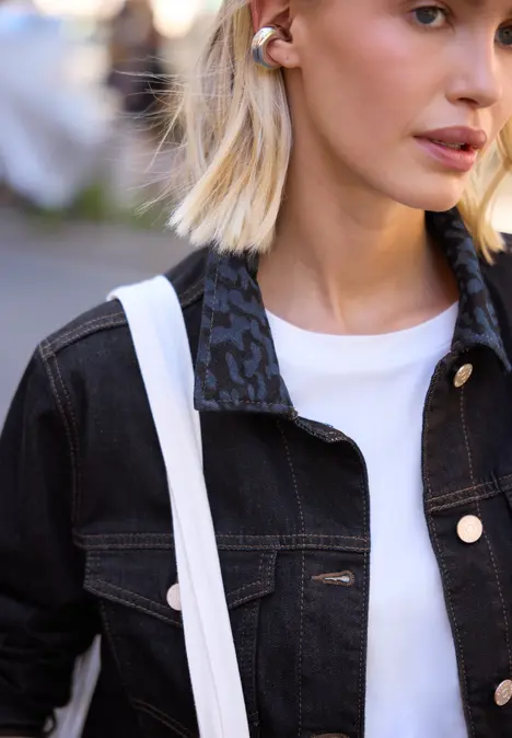 Junge Frau mit blonden Haaren, schwarzer Jeansjacke, weißem T-Shirt und großer weißer Tasche.