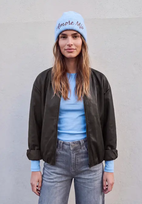 Junge Frau in grauen Jeans, hellblauem Pullover, dunkler Jacke und hellblauer Mütze vor schlichtem Hintergrund.