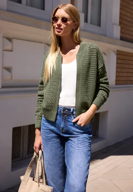 Junge Frau in grüner Strickjacke, weißem Top, Jeans und Sonnenbrille trägt eine Tasche in sonniger Straße.