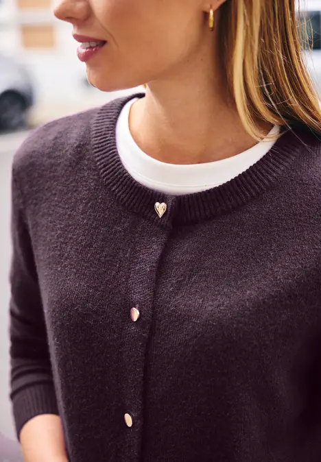 Frau trägt dunkelviolette Strickjacke mit goldenen Herzknöpfen über weißem Shirt.