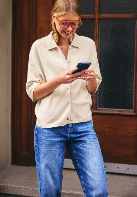 Eine Frau in Jeans und beigem Hemd steht vor einer Tür und schaut lächelnd auf ihr Smartphone.
