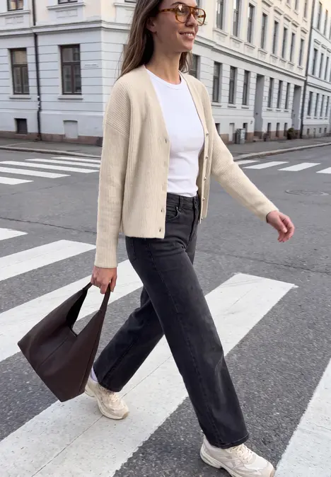 Frau mit Brille in Cardigan, weißem Shirt und dunklen Jeans trägt Tasche und überquert Zebrastreifen.