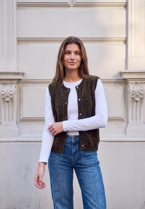 Frau lehnt lächelnd an einer Fassade, trägt weißes Shirt, Weste und Jeans.