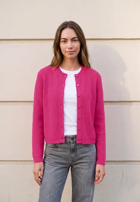 Eine Frau steht vor einer hellen Wand, trägt eine pinke Strickjacke über weißem Shirt und Jeans.
