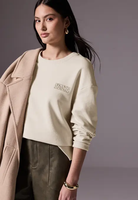 Frau mit beigem Pullover, Mantel über der Schulter und goldenen Accessoires.