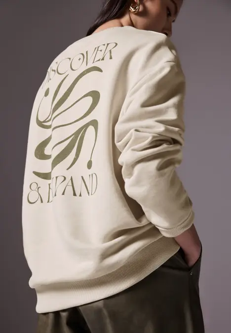 Frau mit beigem Pullover, Rückseite mit Schrift und Grafik.